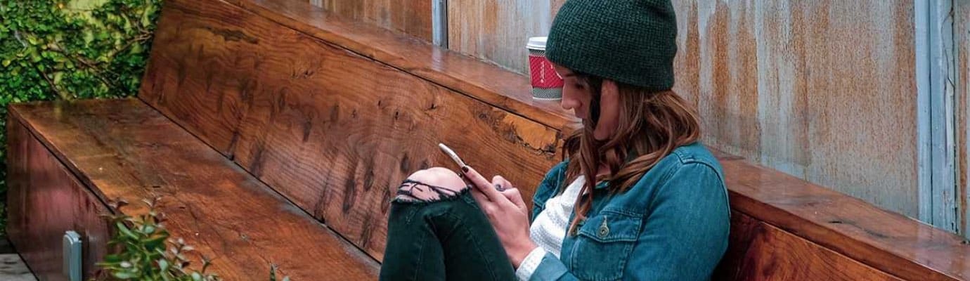 Junge Frau sitzt auf Holzbank und schaut in ihr Handy