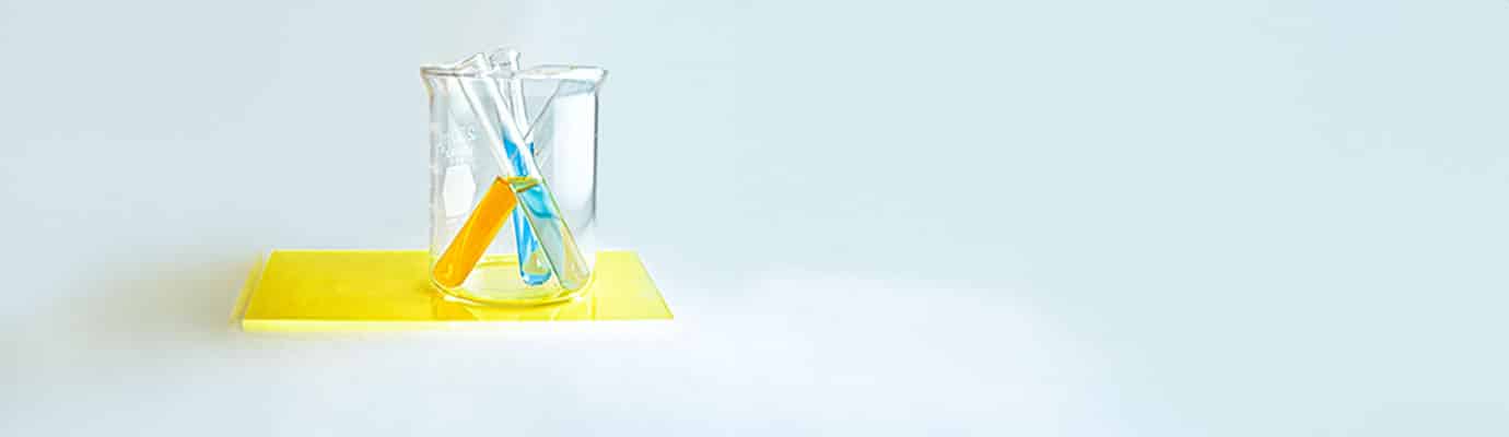Ein Labor-Reagenzglas mit drei bunten Glasröhrchen steht auf gelben Tablett auf hellblauem Grund