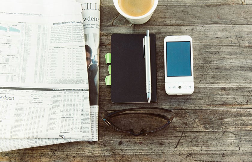 Zeitung, Notizbuch mit Stift, Mobiltelefon, Kaffeebecher und Sonnenbrill auf einem Holztisch