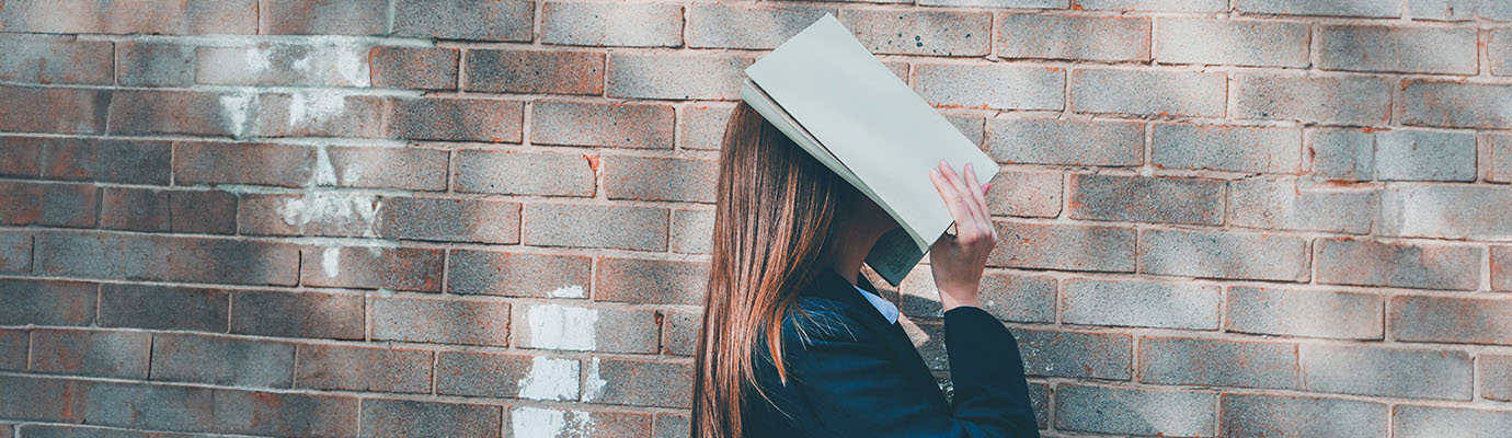 young woman in front of a brick wall holds an open A4 booklet in front of her face
