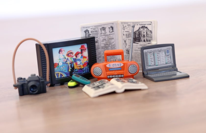 Spielfiguren und Playmobil-Gegenstände auf einem Haufen: Radio, Laptop, Fernseher, Kamera, Buch und Zeitung.