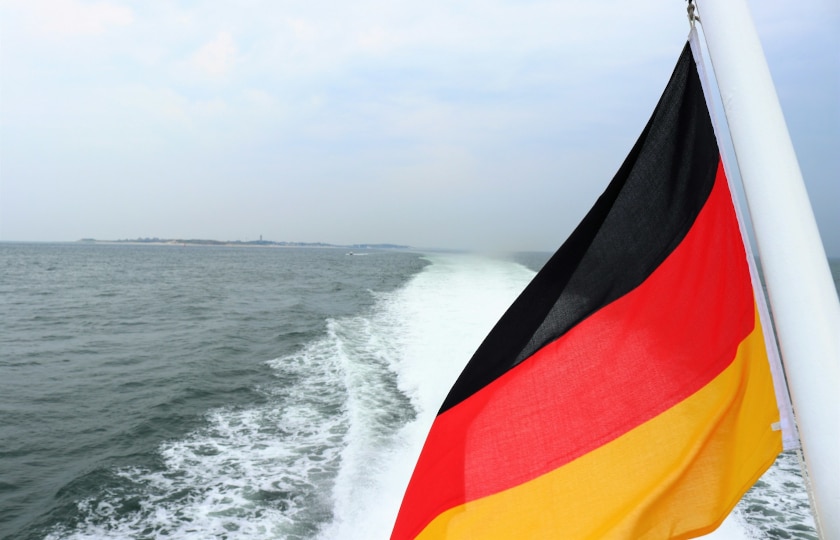 Deutschlandflagge weht an Schiffsheck. Dahinter Fahrwasser und weites Meer