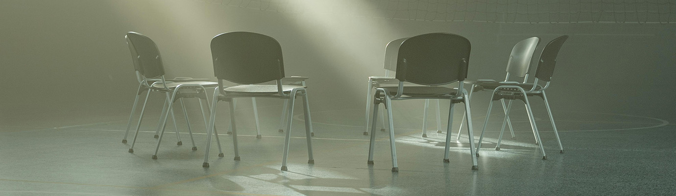A beam of light falls on an empty circle of chairs from the high window.