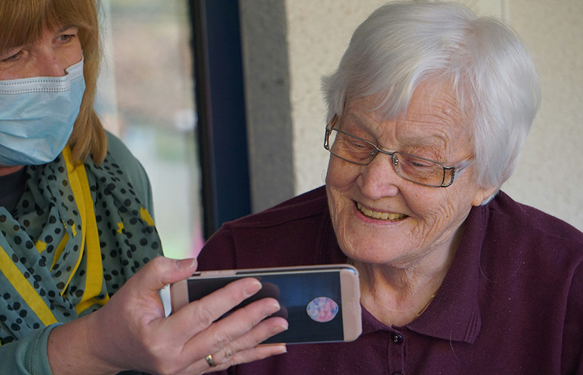 Eine Frau mit Maske zeigt einer Seniorin etwas auf dem Samrtphone