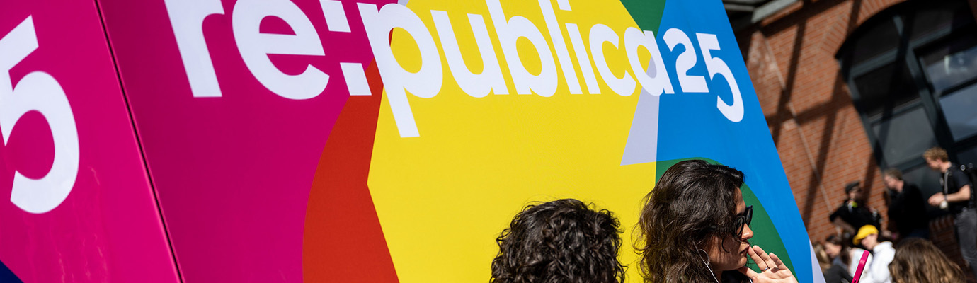 Two young women sit looking at their cell phones in front of a large red, yellow, green, and blue banner with the RePublica25 logo.