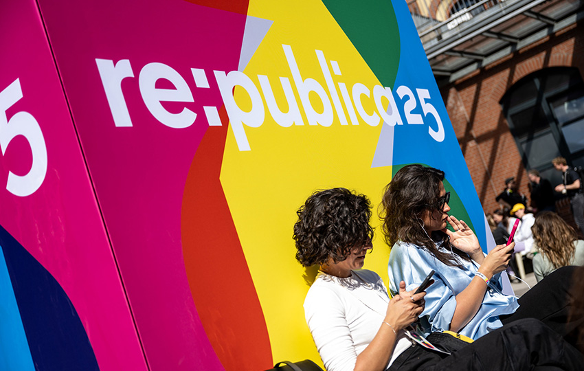 zwei junge Frauen sitzen auf ihr Handy blickend vor einem großen rot-gelb-grün-blauen Banner mit RePublica25-Logo