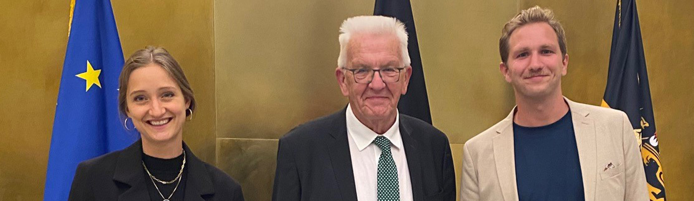 Leonie Alatassi, Minister President Winfried Kretschmann, and Kilian Hampel in front of a golden wall with the European flag