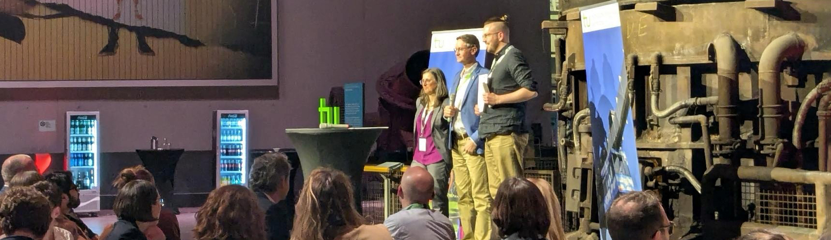 Three people are standing on a stage in front of an audience in a room with an industrial feel