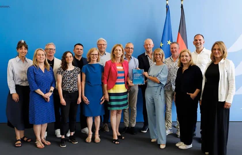 Expertenkommission mit Bundesministerin Prien vor einer blauen Wand mit den Flaggen der Europäischen Union und Deutschlands