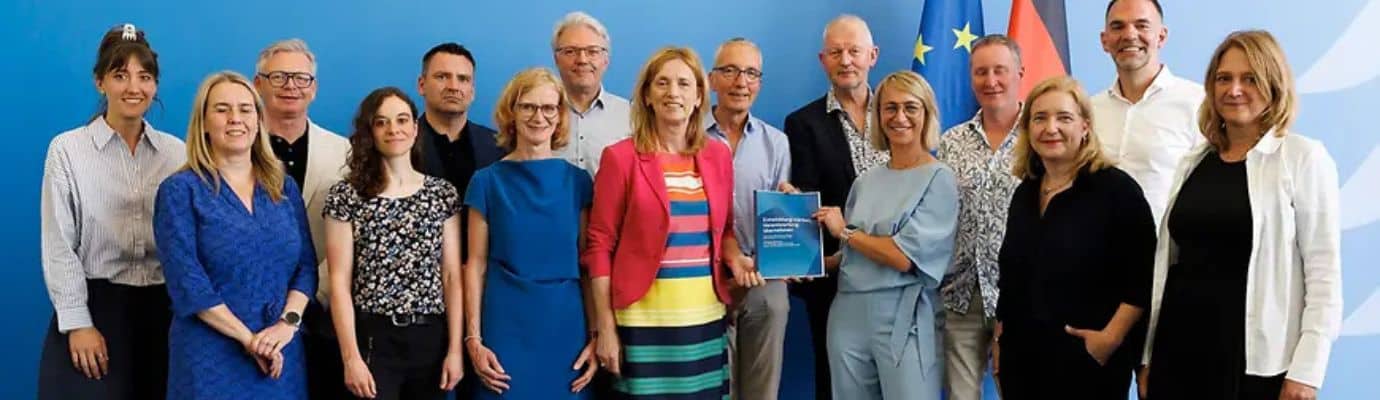 A commission of experts with Federal Minister Prien in front of a blue wall displaying the flags of the European Union and Germany