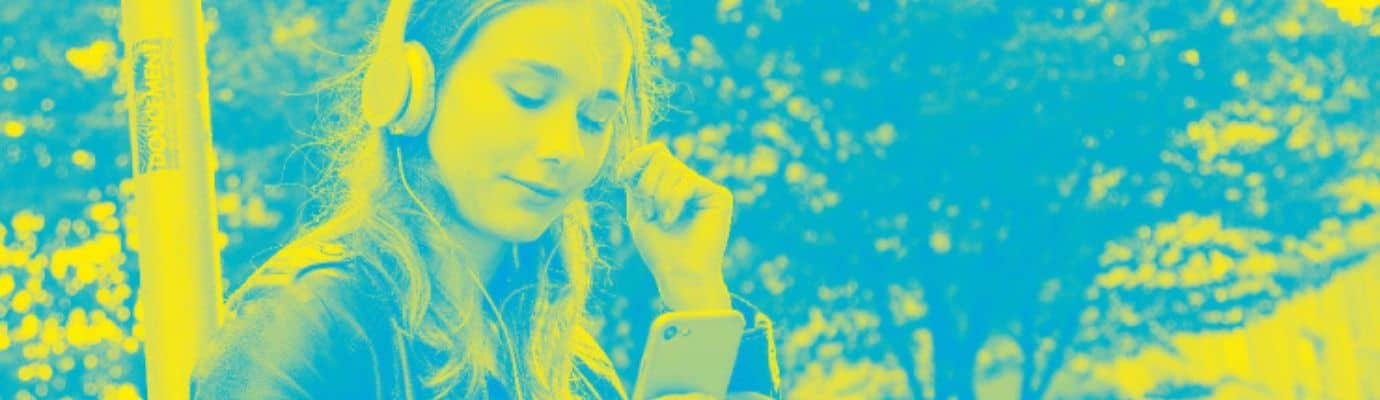 A girl is sitting with headphones on, leaning against a pole and looking at her cell phone.