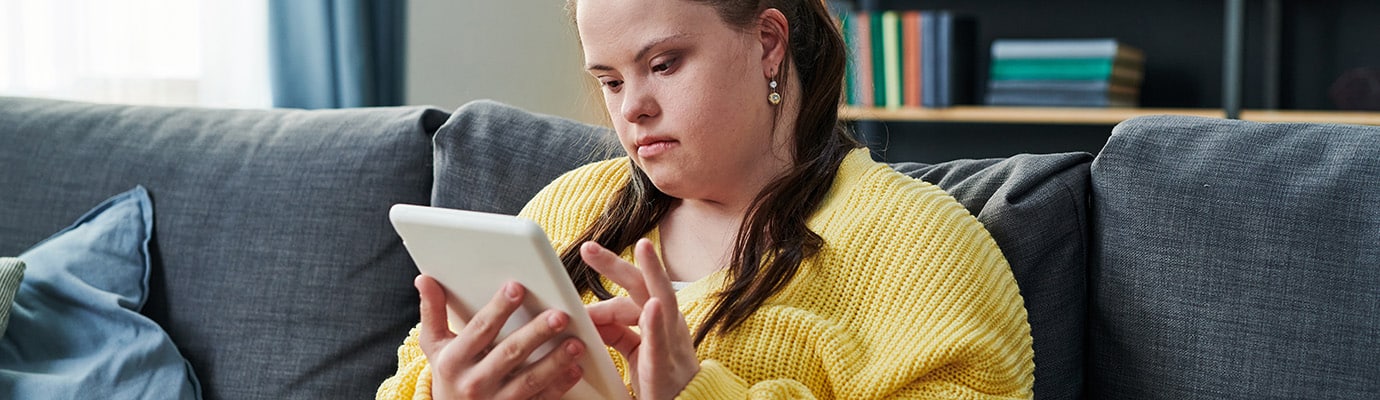 Portrait of modern girl with Down syndrome sitting on couch in living room at home reading book on digital tablet