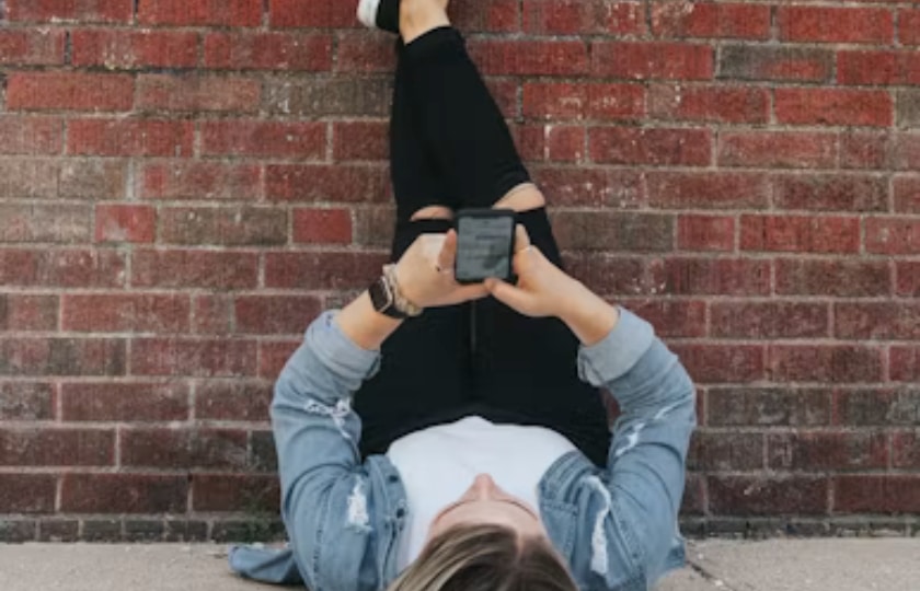 Eine Junge Frau liegt, die Beine hoch an einer Klinkerwand, und schaut auf Ihr Handy