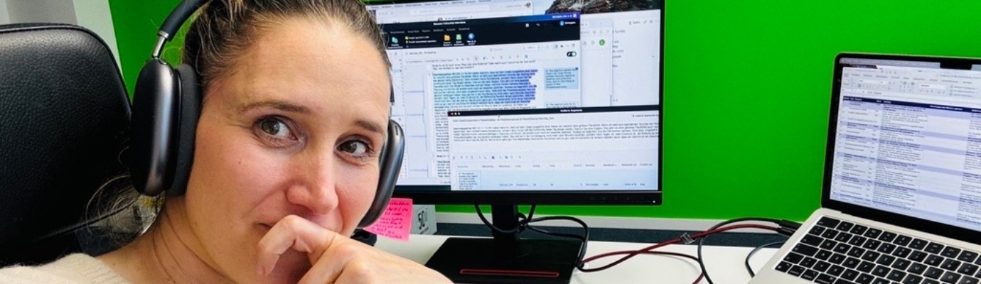Svea Eckert, wearing headphones, looks thoughtfully into the camera; behind her are her work laptop and a screen displaying text