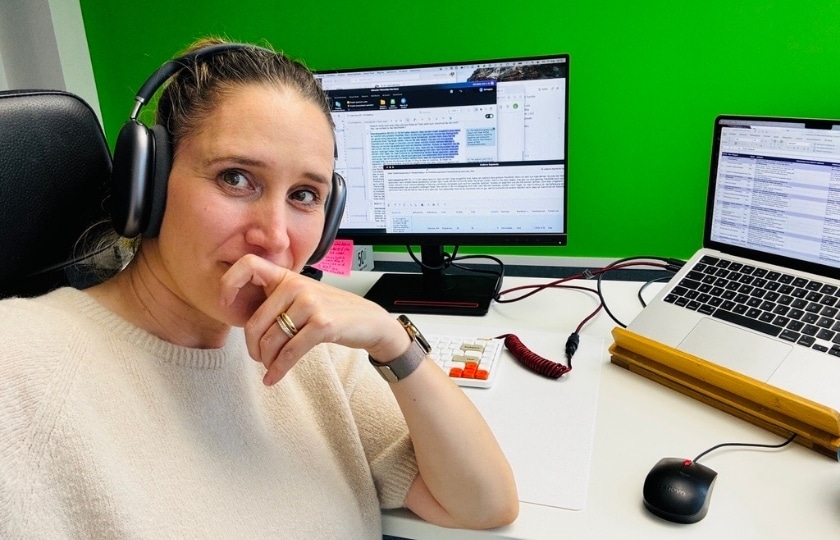 Svea Eckert mit Kopfhörern schaut nachdenklich in die Kamera, hinter ihr sind Arbeits-Laptop und Bildschirm mit Text zu sehen
