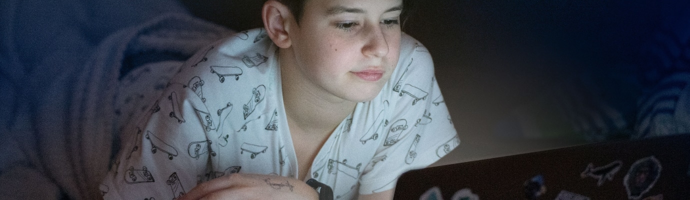Jugendlicher liegt bäuchlings in eine Decke gewickelt und schaut bei schwachem Licht in einen leuchtenden Laptop