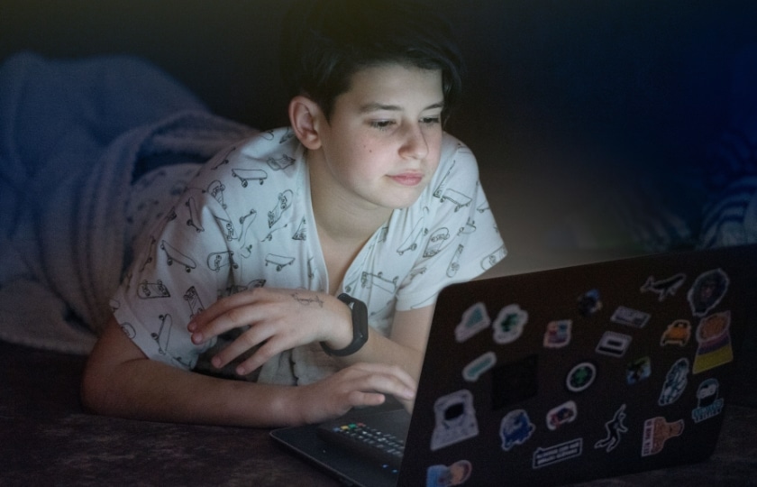 Jugendlicher liegt bäuchlings in eine Decke gewickelt und schaut bei schwachem Licht in einen leuchtenden Laptop